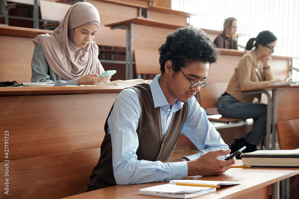 Poster mixed-race guy and other college students in casualwear sitting by wooden desks in lecture hall at l