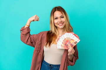 Teenager blonde girl taking a lot of Euros over isolated blue background doing strong gesture