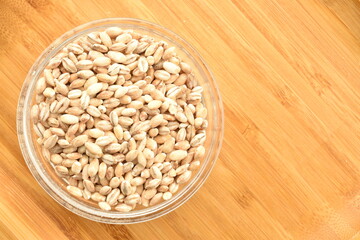 Organic uncooked pearl barley in a glass dish on a bamboo tray, close-up, top view.