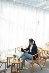 Pretty young female entrepreneur resting in cafe, drinking coffee and reading reports and documents on tablet computer