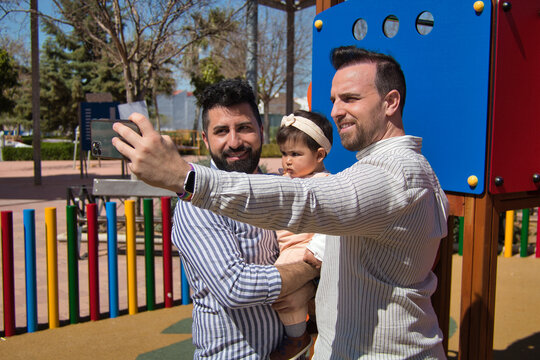 Gay married couple taking a selfie with their cell phone with their daughter in their arms in a playground.