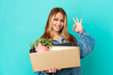 Teenager blonde girl making a move while picking up a box full of things showing ok sign with fingers