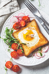 Fried egg in bread toast with tomatoes, radishes and sprouts on white plate, gray background, top view. Healthy breakfast concept.