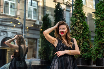 Young brunette girl in elegant black evening dress posign near at front of glass building