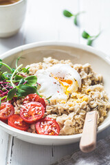 Savory oatmeal with poached egg, tomatoes, cheese and sprouts in white bowl, close up. Healthy breakfast concept.