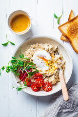 Savory oatmeal with poached egg, tomatoes, cheese and sprouts in white bowl, top view. Healthy...