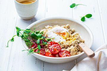 Savory oatmeal with poached egg, tomatoes, cheese and sprouts in white bowl. Healthy breakfast concept.