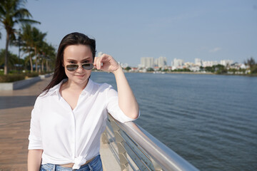 Portrait of young beautiful woman taking off sunglasses and smiling at camera when standing on river bank