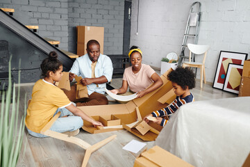 Young African family of sitting on the floor of large living-room while unpacking and taking out furniture parts from cardboard box