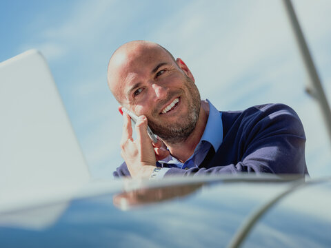 Businessman talking on the phone while using the laptop supported by a car and smiling expression