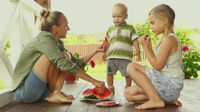 A happy mom smiling while two children eating watermelon on summer day.