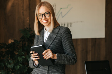 A businesswoman holds a notebook in her hands stands and smiles in the office while planning new projects for her company