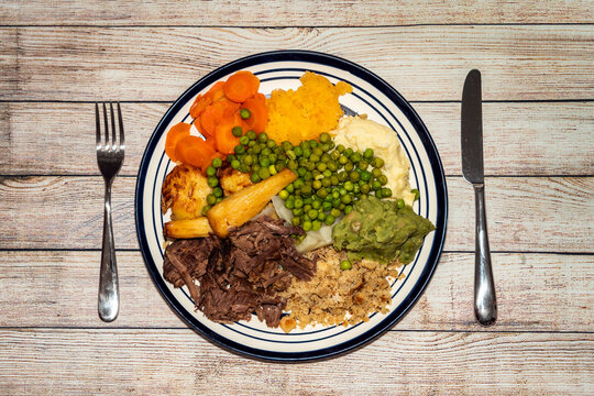 Sunday Dinner Of Slow Cooked Shredded Pulled Beef On A Plate With A Knife And Fork And Plenty Of Vegetables Such As Peas, Mushy Peas, Parsnips, Roast Potatoes And Carrots, Stock Photo Image