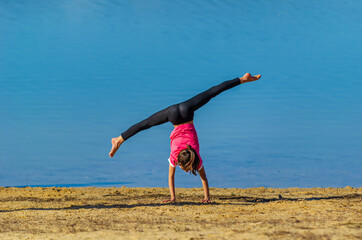 7 years old girl with pink shirt and sportswear doing a cartwheel at the beach during spring with blue water as background. Palencia, Spain - Powered by Adobe