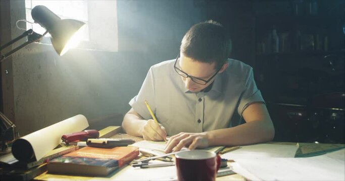 Young man in shirt and glasses working hard in his garage, studying and writing things down.