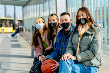 Group of happy four youth people wearing protective facial masks sitting next to other in downtown at bus or tram stop. Urban lifestyle new rules concept.