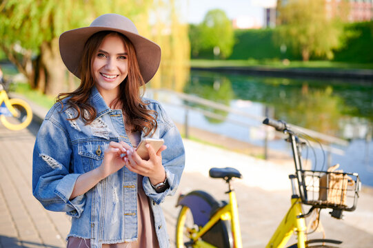 Young fashionable woman in hat and denim jacket use smartphone in public city park in front of yellow rental bike
