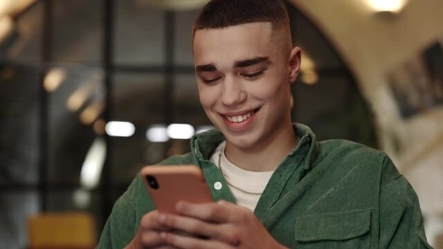 Close up view of young man with stylish haircut using smartphone while sitting in chair. Handsome male person in casual clothes laughing while scrolling social media news feed in cafe.