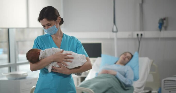 Nurse in medical mask holding newborn baby with mother resting in bed on background