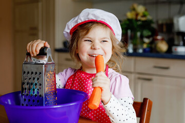 Cute little preschool girl grate carrots for baking carrot cake or cookies. Happy toddler child in...
