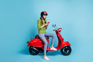 Full length photo portrait of excited girl holding phone in two hands sitting on red scooter isolated on pastel blue colored background © deagreez