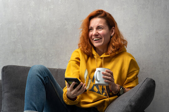 Cheerful red-haired girl with cup and telephone in hands on sofa. Chatting with friends