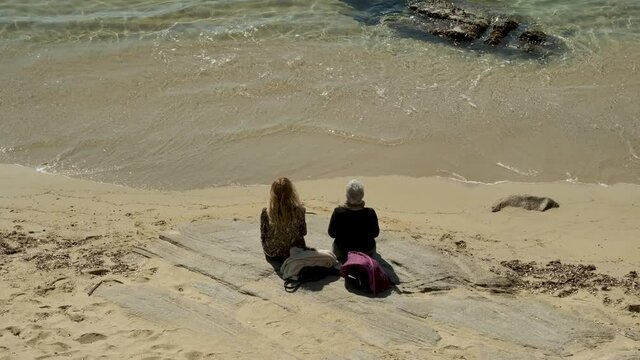 Mother and daughter or friends meditate looking at sea water. Elderly couple sitting near each other on sandy beach on sunny day. Romantic relationships in older people. Vacation, holidays in sea.