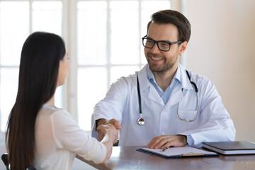 Smiling young male GP or therapist in white medical uniform handshake female patient close health insurance deal in hospital. Happy man doctor shake hand of client get acquainted or greet in clinic.