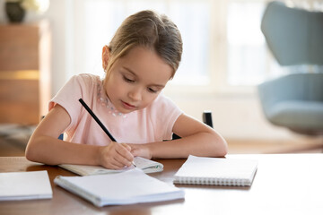 Smart small 7s Caucasian girl child sit at table at home write in exercise book prepare homework for school. Little kid handwrite in notebook do task assignment. Learning, education concept.