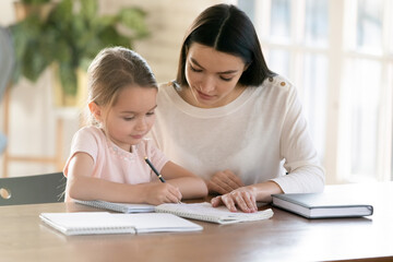 Loving young Caucasian mother and little daughter write in exercise book do homework. Caring mom...