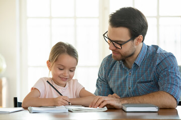 Caring young father help small daughter do homework write in exercise book at home. Loving...
