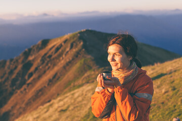 Close-up portrait of young girl drinking hot tea from steel mug in background of sunset. Woman traveler holds mug with coffee and looks at beautiful sunny mountain. Scenic photo, copy space