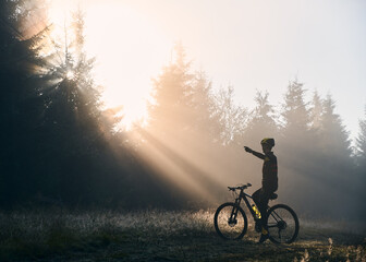 Back view of young man sitting on bicycle and pointing finger at beautiful morning sunlight behind trees. Male bicyclist sitting on mountain bike in morning forest.