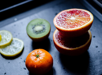 Mix of fresh tropical fruits including blood orange, kiwi, clementine and lemon slices on back worktop covered with water drops.