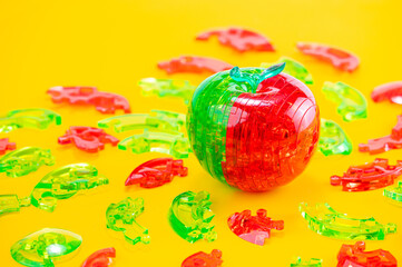Assembled red-green plastic puzzle in the shape of an apple, surrounded by pieces of plastic puzzle, on the yellow background.