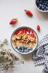 Colorful food flat lay: lush bowl of muesli nuts mix, cereal, fresh strawberries, blueberries, banana slices and yogurt on white with wax flowers and rustic table cloth. Healthy breakfast. Top view.