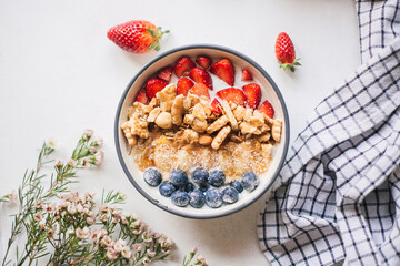 Colorful food flat lay: lush bowl of muesli nuts mix, cereal, fresh strawberries, blueberries, banana slices and yogurt on white with wax flowers and rustic table cloth. Healthy breakfast. Top view.