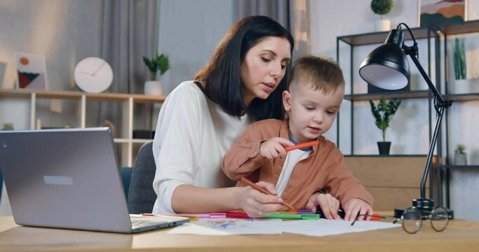 Portrait of pretty loving caring 30-aged woman which holding small attantive son on knees and helping him to draw beautiful picture