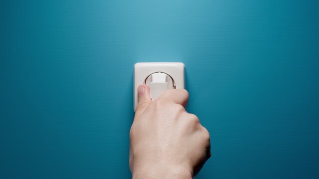 Human hand inserts a plug into an electrical outlet on a blue wall