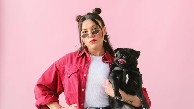 Young stylish unusual woman posing holding small black pug dog in her arms on pink background. Trendy generation.