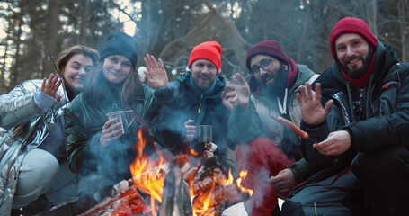 Happy young friends sitting by campfire in winter forest and waving hands at camera