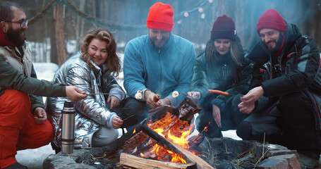 Happy campers sitting by fire and roasting marshmallows and sausages on stick in winter forest