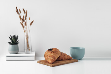 Croissant and a mug on a wooden stand, a vase and a pot with a plant on the books, white background. Eco-friendly materials in interior decor, minimalism. Copy space, mock up