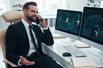 Smiling young man in shirt and tie analyzing data on the stock market and talking on the phone while working in the office