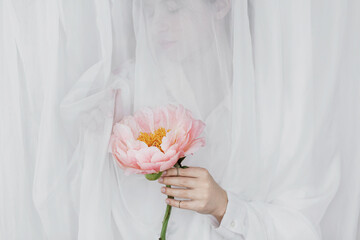 Sensual beautiful woman behind soft white fabric with pink peony in hands. Tender bridal morning