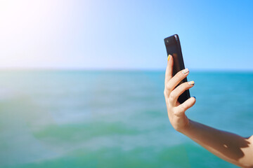 Mobile phone in woman's hand. Roaming and communication concept. Girl holds smartphone on sea background. Sunny summer day.