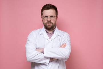 Serious doctor in a white coat and glasses looks sternly and stands with crossed arms. Pink background.