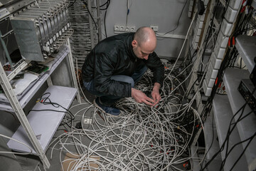 A technician sits on wires in a ransacked server room. The man reconnects at the abandoned datacenter. An engineer repairs equipment of an old TV station
