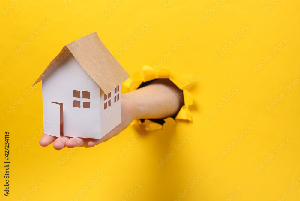 Wall mural female hand holds house model through torn hole yellow paper. concept art. minimalism