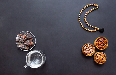 Ramadan Kareem and iftar muslim food and glass of clear water, modern holiday concept. Wooden bowls with nuts and dried fruits and the moon made of beads for rosary with tassel on a black background 